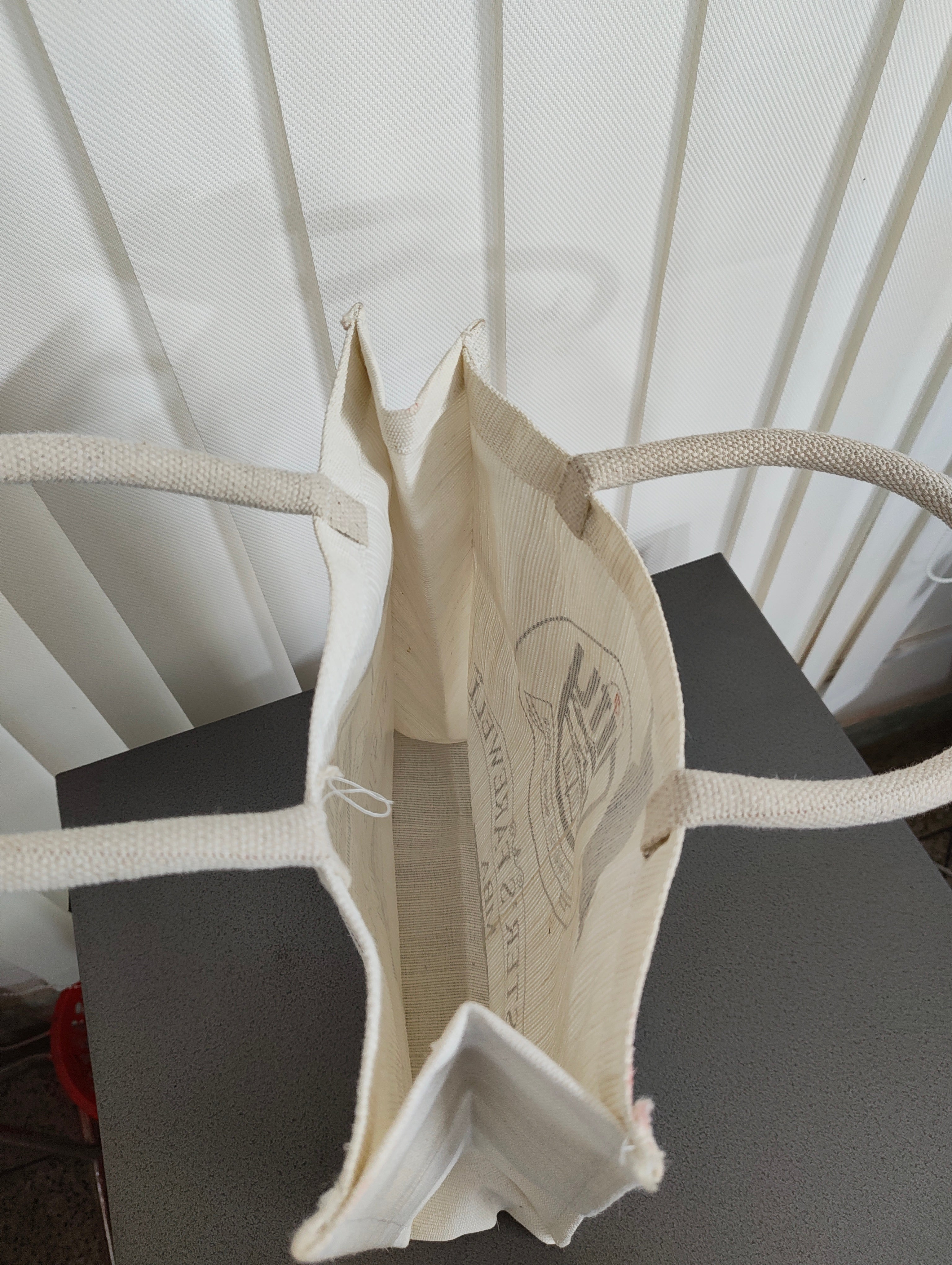 An off-white canvas tote bag with thick rope handles, shown from a top-down perspective resting on a dark gray surface in front of a white slatted screen.