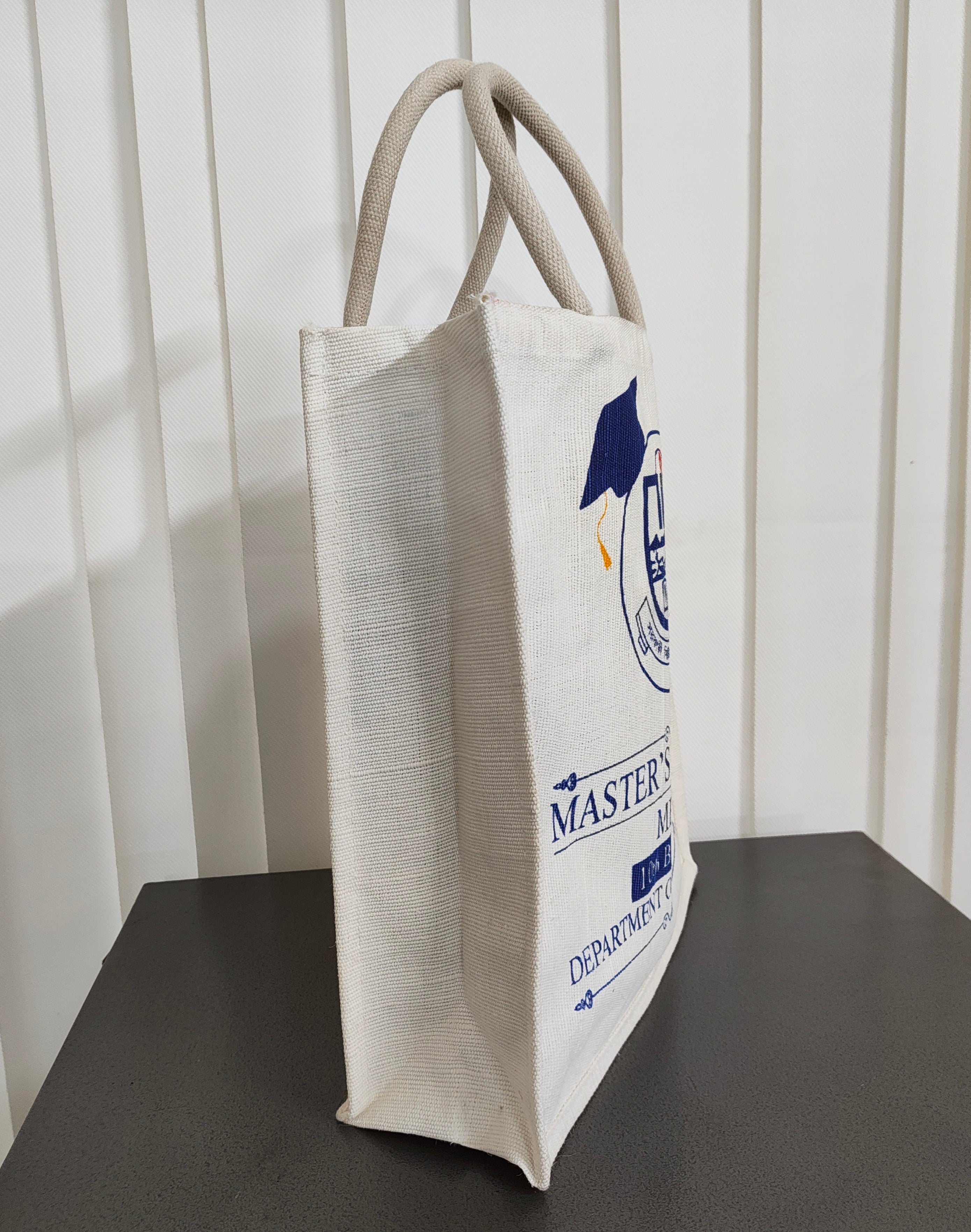 An off-white canvas tote bag with beige rope handles, featuring a navy blue and white 'Master of Arts Department of Art' graphic, standing on a dark gray surface against a white paneled wall.