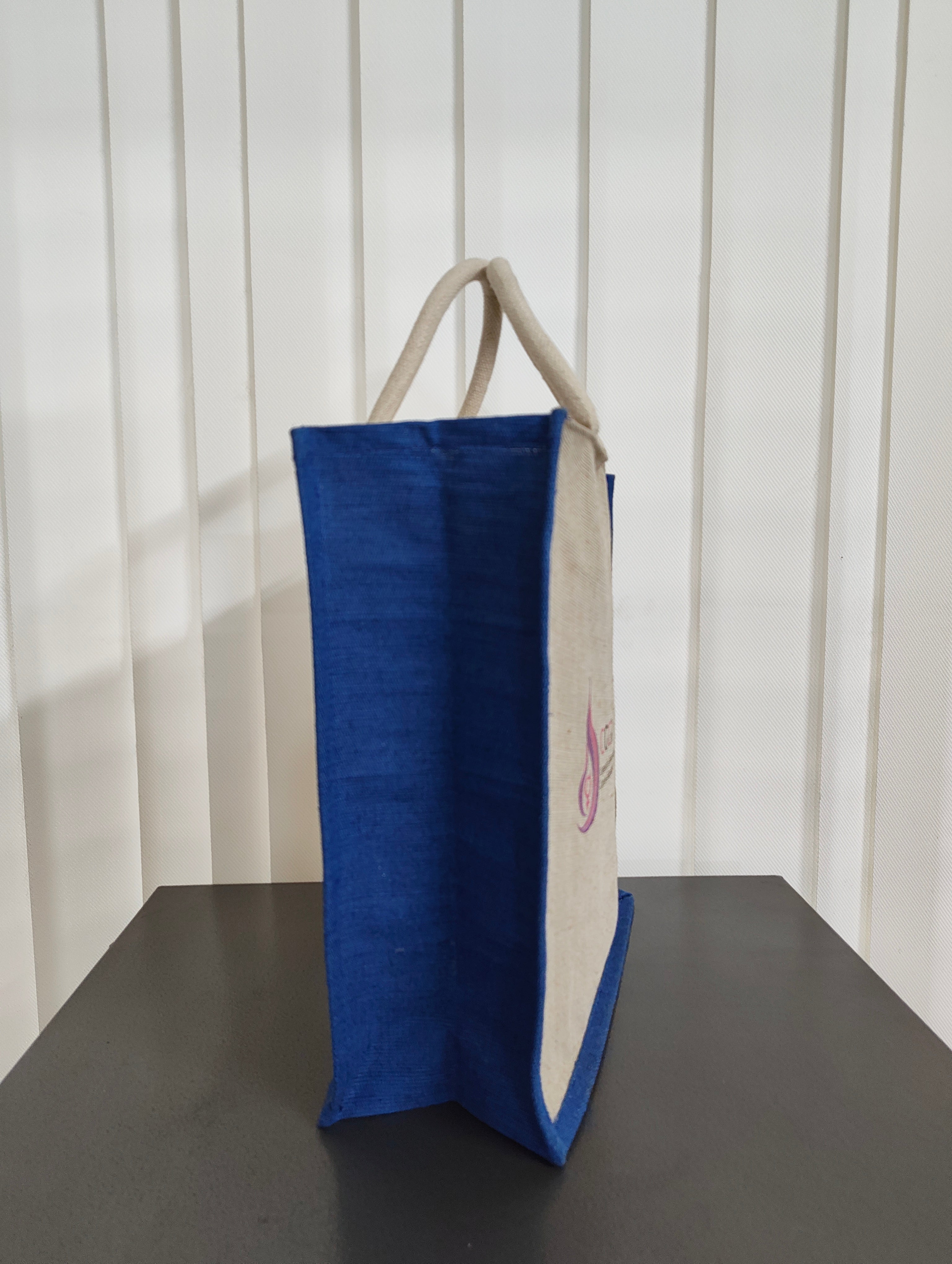 A blue and beige canvas tote bag with beige shoulder straps, standing on a dark surface against a white slatted wall.