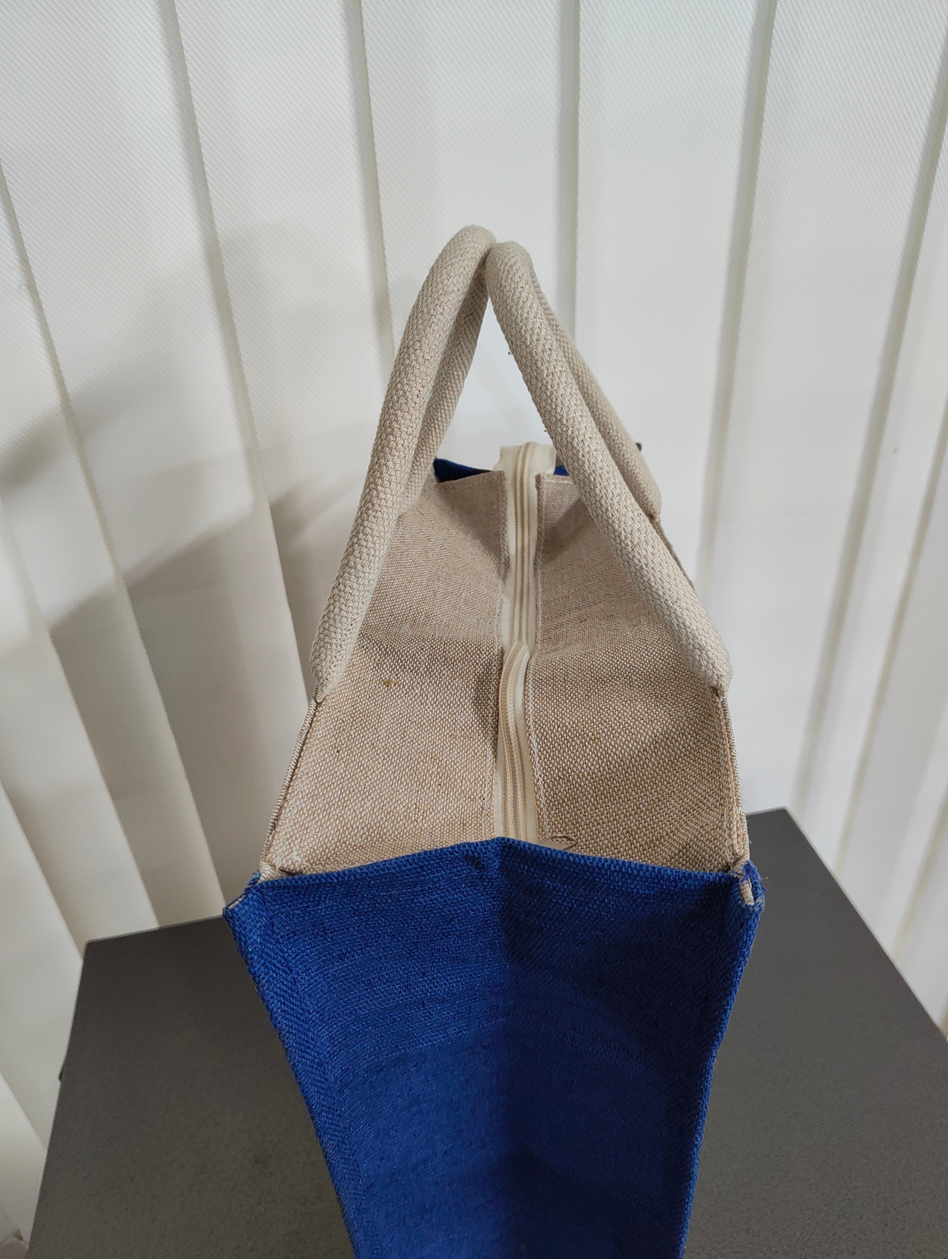 A canvas tote bag with a natural beige exterior, cream zipper, and a vibrant blue lower half, shown standing on a dark surface against a white paneled background.