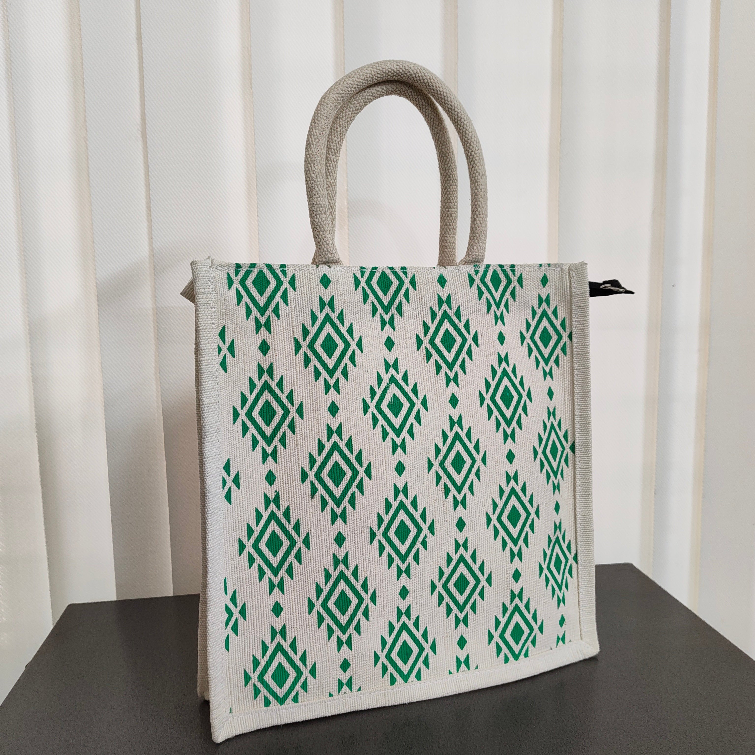A white canvas tote bag with a green geometric diamond pattern and thick light gray handles, standing on a dark surface against a white slatted background.