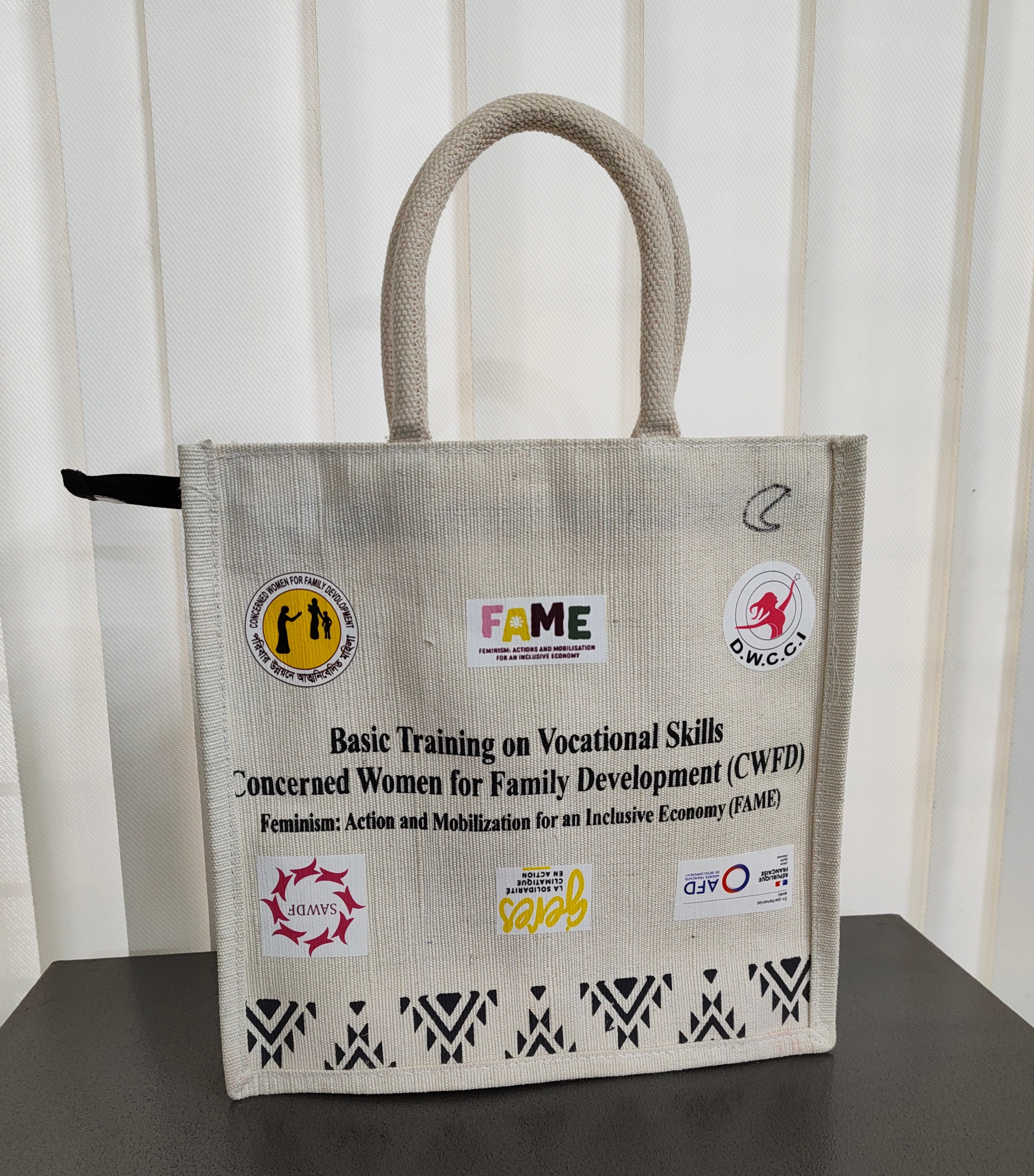 A beige jute tote bag with dual top handles, featuring printed organizational logos and text including FAME, CMCC, and SWAB, displayed on a dark surface against a white paneled background.
