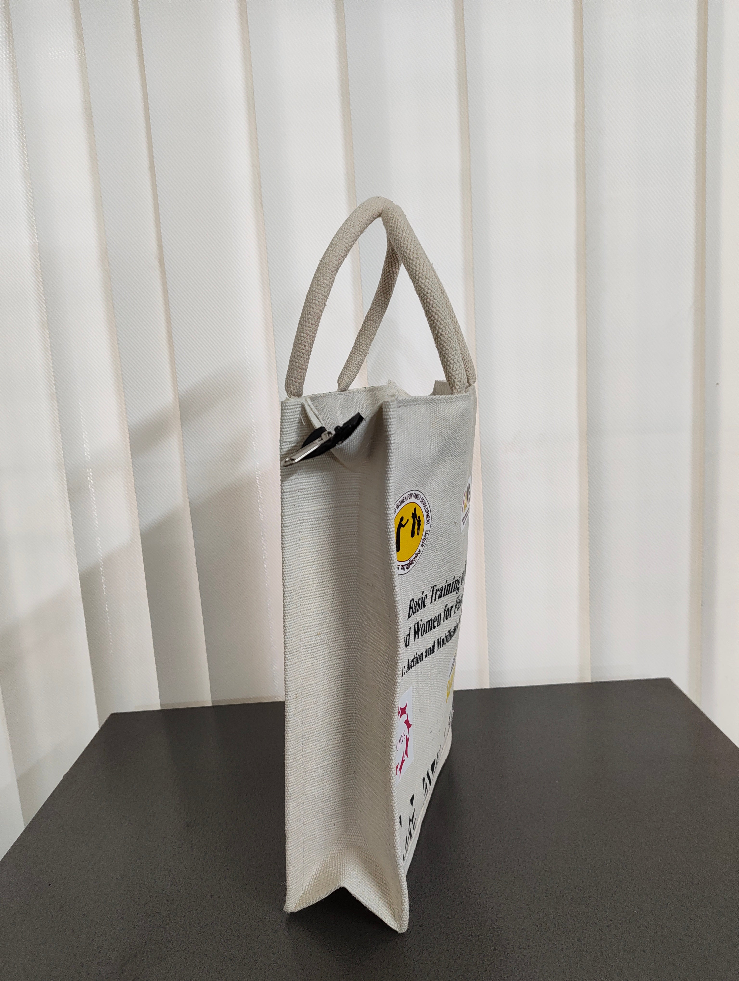 A light beige canvas tote bag with dual top handles, standing upright on a dark tabletop in front of white vertical blinds. The bag features a yellow circular logo and black text on the side.