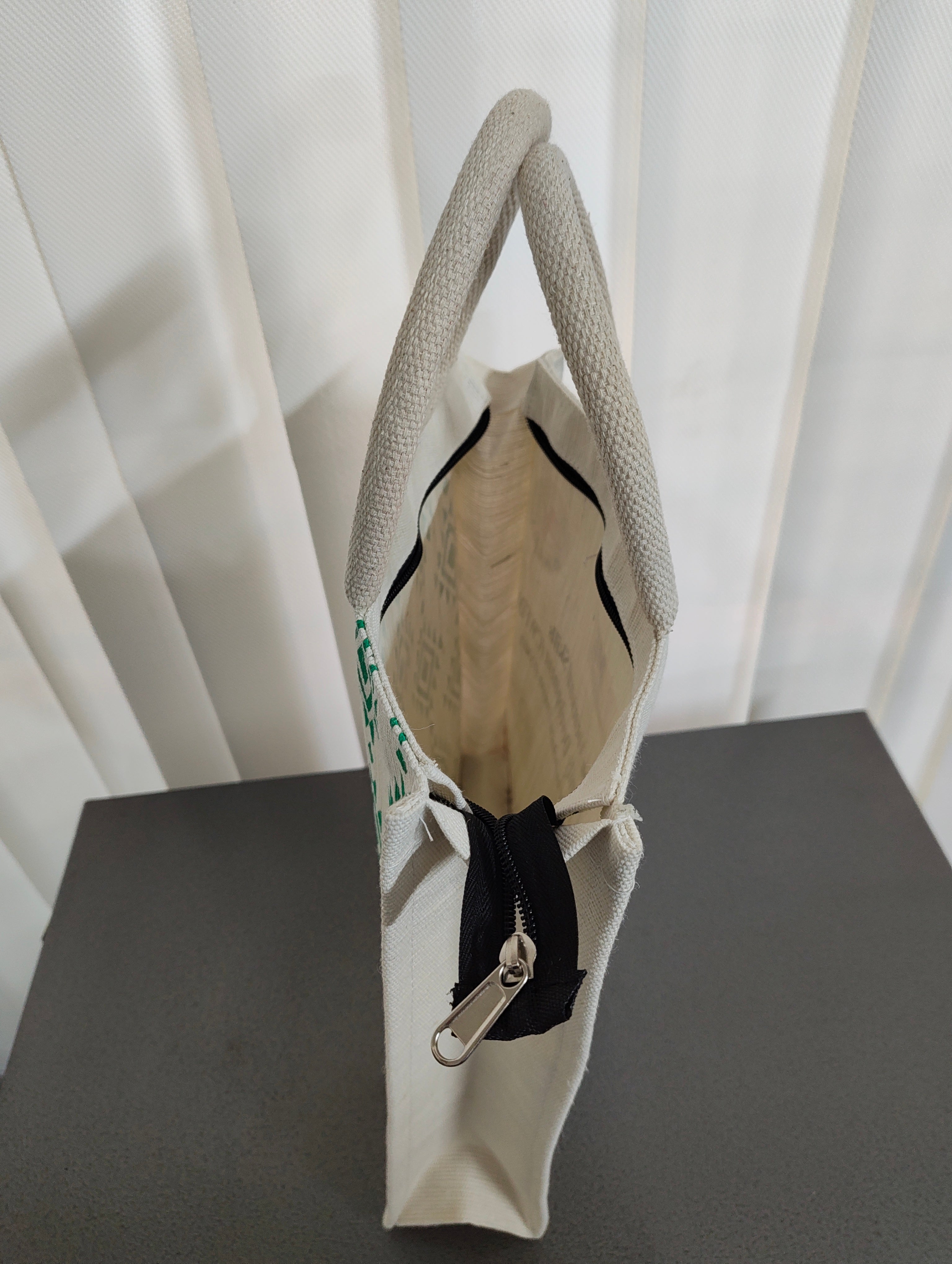 A cream canvas tote bag with textured gray handles, a black zipper closure, and a silver metal pull, shown standing on a dark gray surface against a white curtain background.