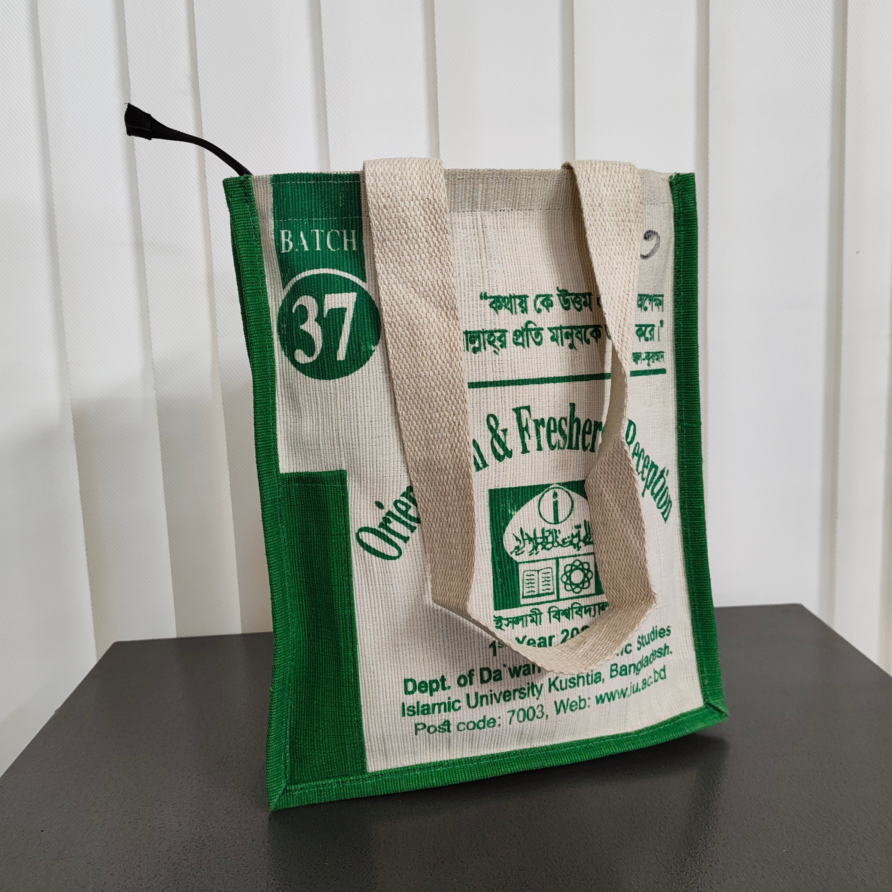 A green and off-white jute tote bag with printed text and logos, standing on a dark surface against a white paneled background.