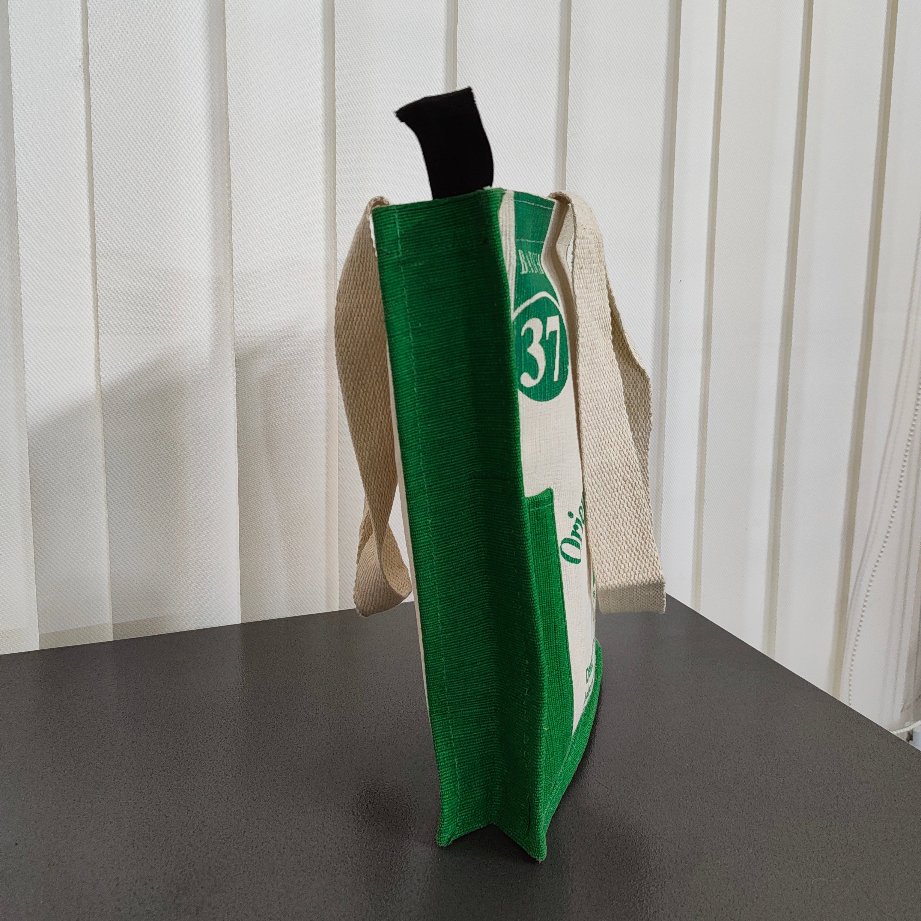 A green and white reusable tote bag with beige canvas shoulder straps, standing upright on a dark tabletop against a white slatted background.
