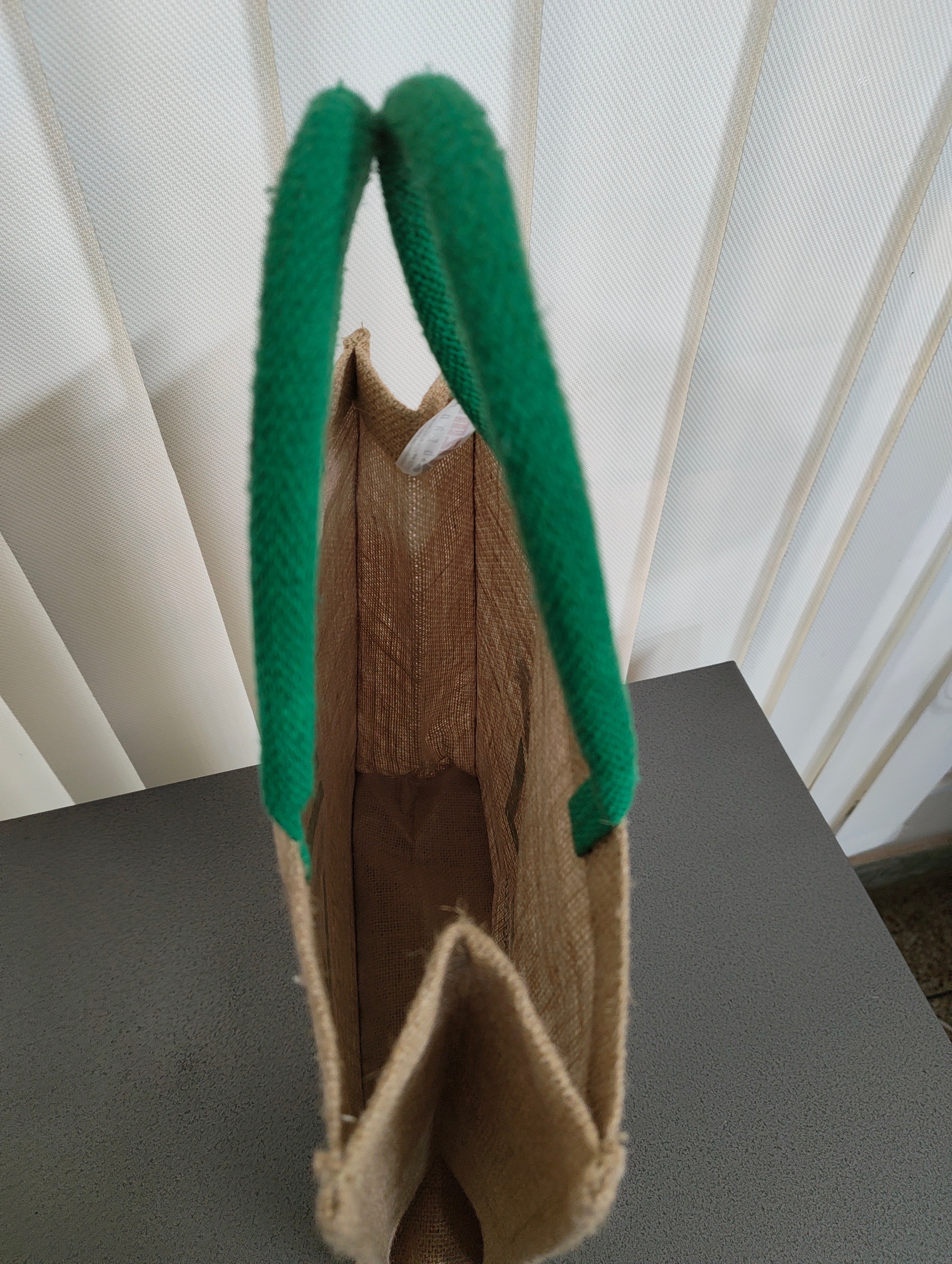 A natural jute tote bag with thick, vibrant green rope handles, shown standing upright on a dark gray surface in front of white vertical blinds.