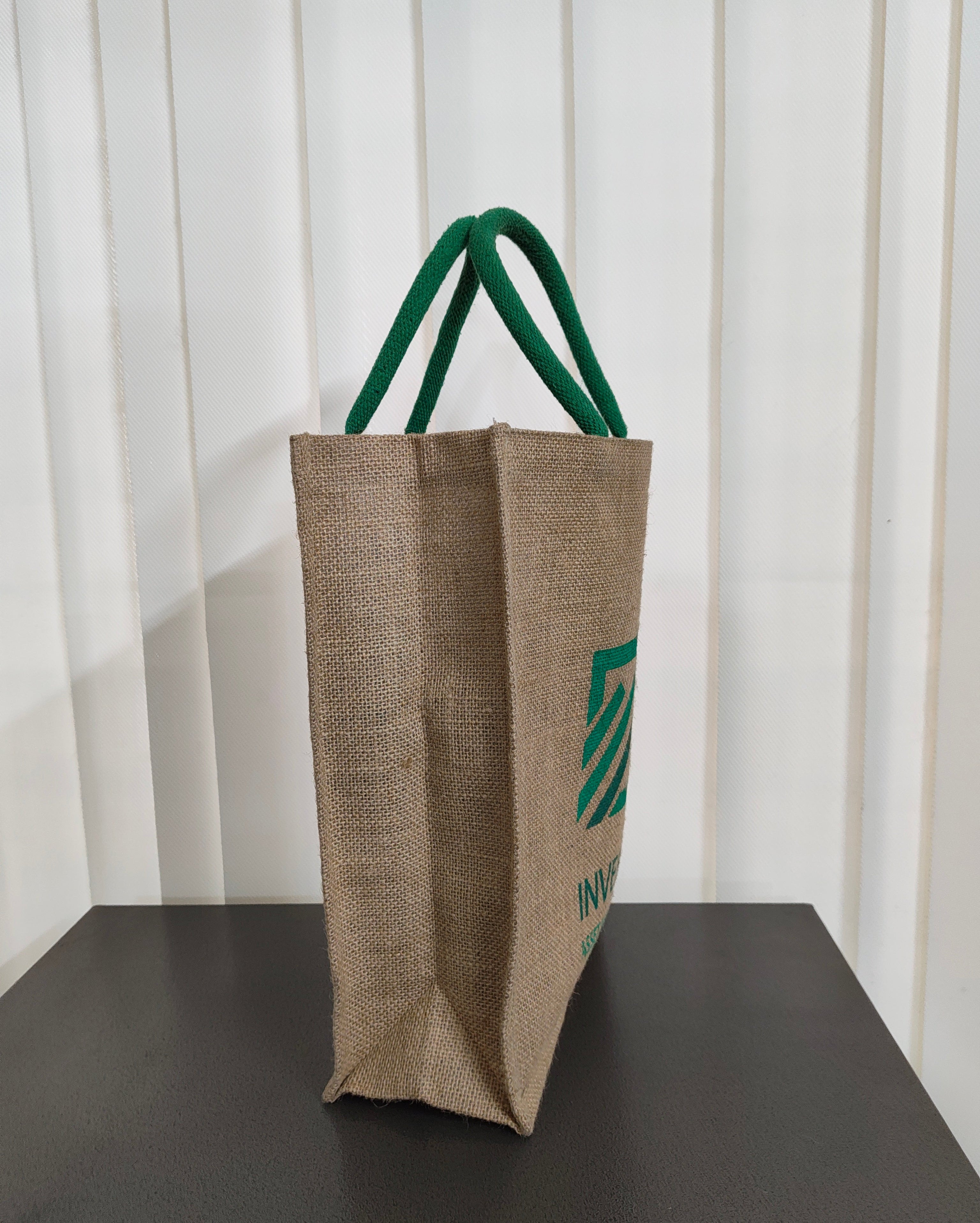 A natural jute tote bag with green fabric handles and a green printed logo, standing on a dark surface against a white paneled background.