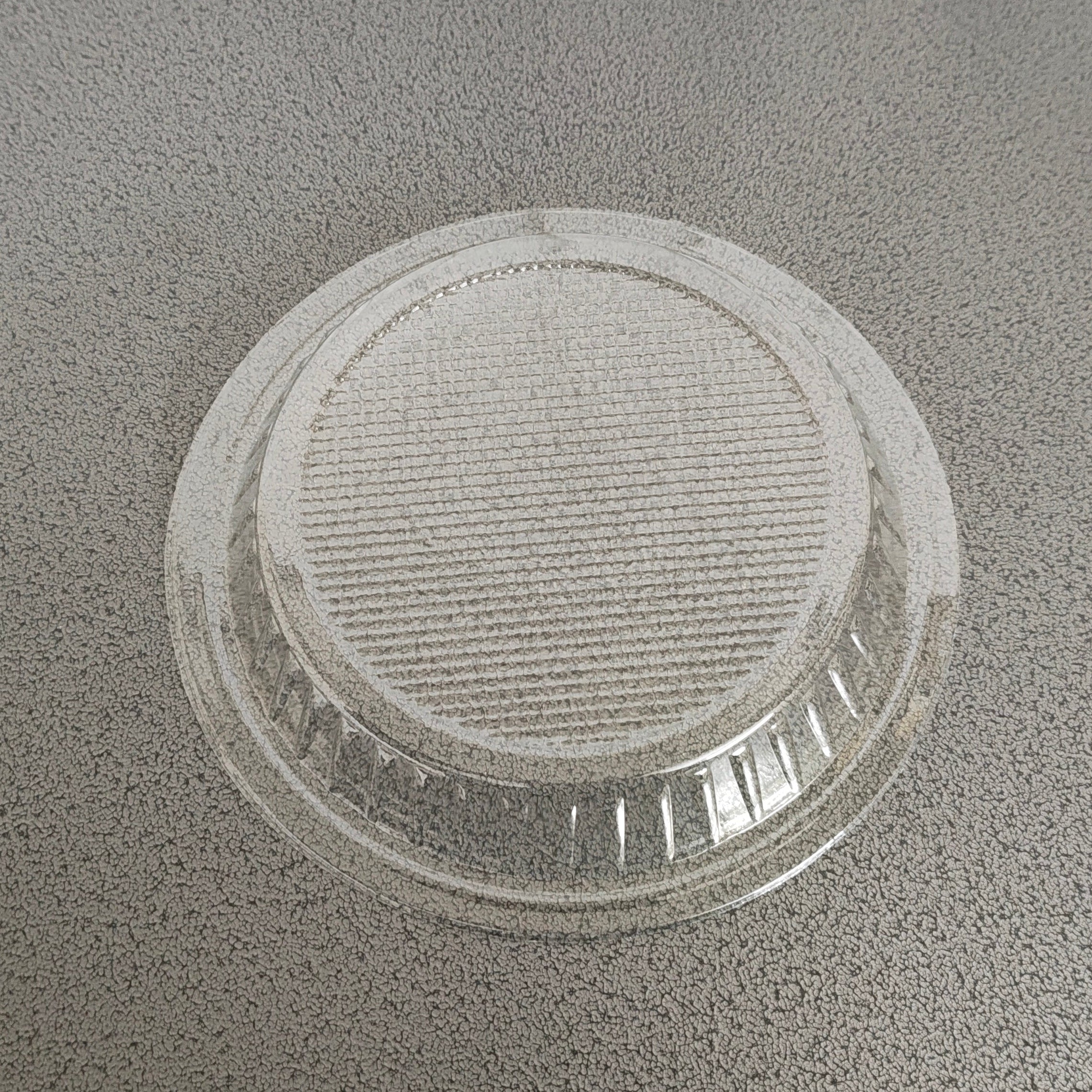 A clear plastic round shallow dish or lid with a ribbed textured top, shown on a speckled gray countertop.