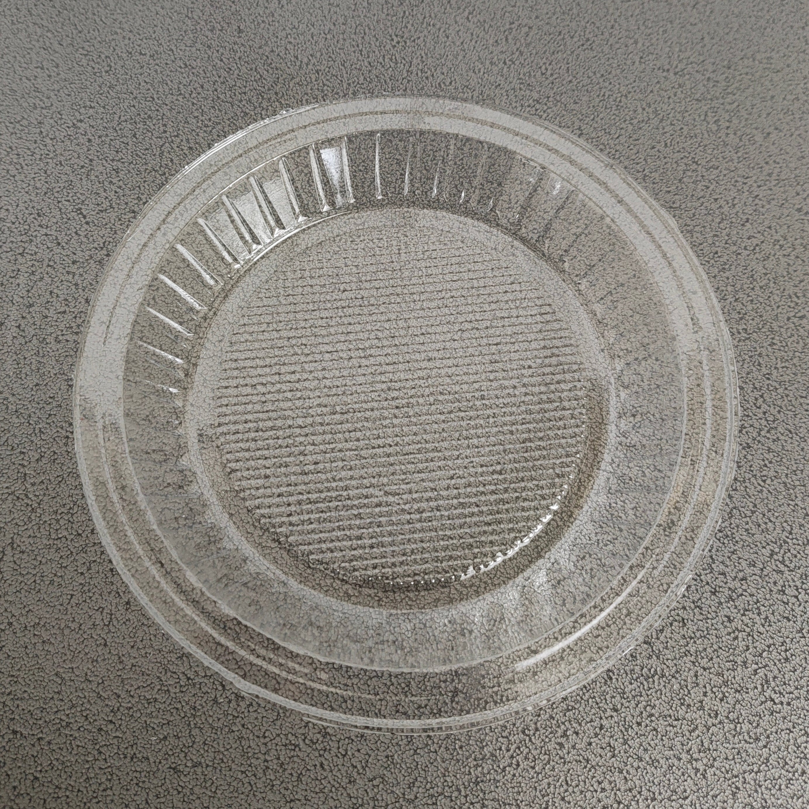 A clear plastic microwave turntable lid or insert tray with a ribbed outer rim and textured center, shown on a speckled gray countertop.