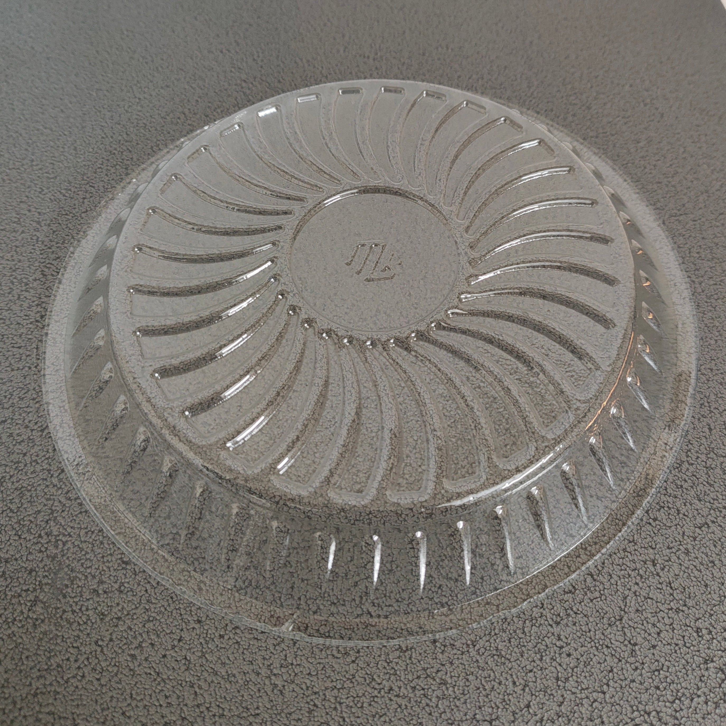 A clear plastic round shallow dish with a ribbed, sunburst-like pattern on the bottom, photographed on a dark gray speckled countertop.