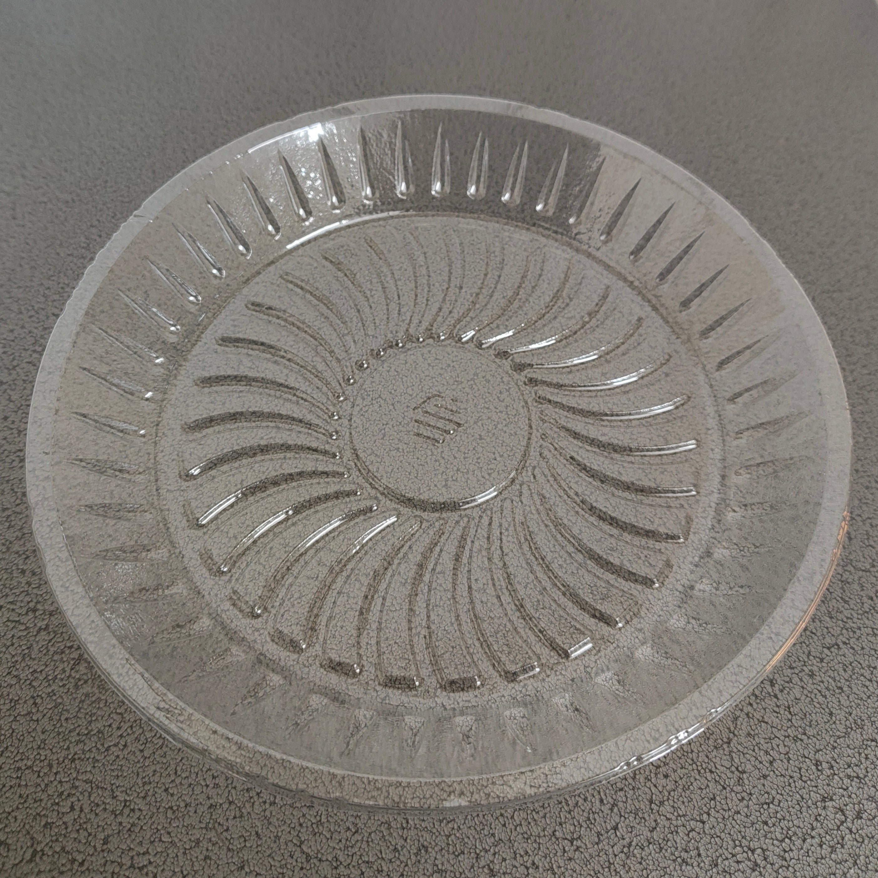 A clear glass plate with a sunburst pattern and a small embossed logo in the center, resting on a dark gray speckled countertop.